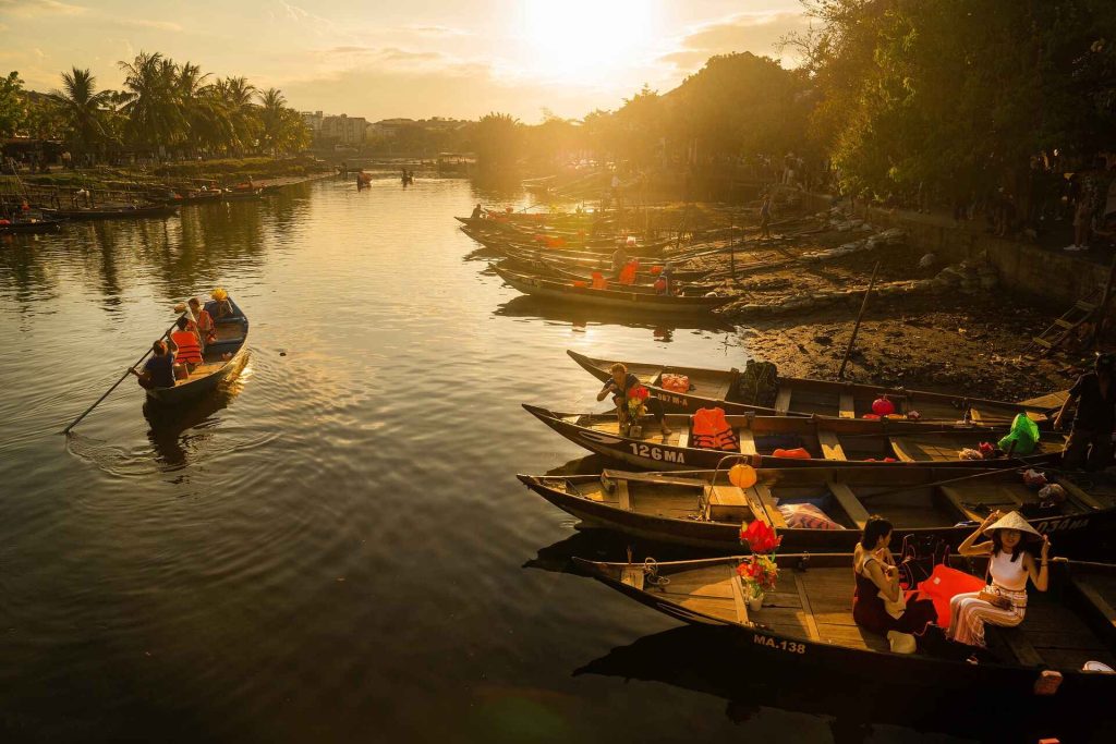 Stunning sunset in Hoi An, Vietnam, with the sky painted in vibrant hues of orange and pink. Silhouettes of traditional Vietnamese buildings and boats on the Thu Bon River reflect in the calm waters, creating a peaceful and romantic atmosphere.